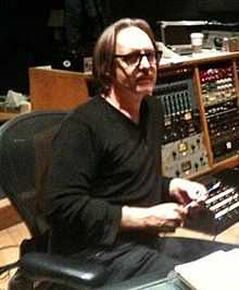 A man wearing glasses and black clothes sits in the control room of a recording studio.