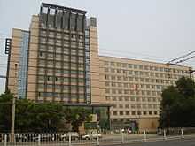 A tall brown modern building with many square glass windows. A front wing on the left is taller than a longer one on the right. There is a metal guardrail in front, at the bottom of the image, and a red flag on a pole in front of the building