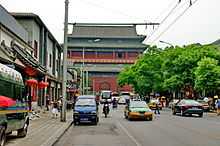 A view up a well-trafficked street to a large dark red building with a Chinese-style roof. On either side are rows of shops. In the foreground is a large metal gantry holding up wires for a trolleybus in the background