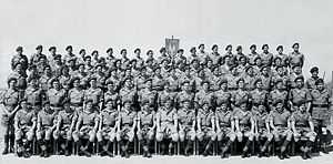 A formative black-and-white photograph of military personnel. The men wear khaki shirts and shorts with long, dark-coloured socks. They all wear dark berets.