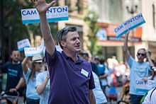 A man in his mid-30s wearing a purple polo shirt waves as people behind him hold up signs with his name, Cam Winton.