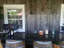 A black marble bar with wine bottles sitting atop oak barrels, with a wooden wall and white-colored window in the background.