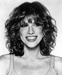 A black and white photo of a blond, curly haired woman wearing a tank top is seen smiling.