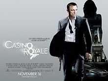 A man in a business suit with a loose tie holding a gun. Behind him is a silhouette of a woman showing a building with a sign reading "Casino Royale" and a dark grey car below the building. At the bottom left of the image is the title "Casino Royale" – both "O"s stand above each other, and below them is a 7 with a trigger and gun barrel – and the credits.