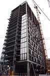 Ground-level view of 19-storey building; the unfinished exterior is white with dark windows, but steel framework is visible at many floor levels