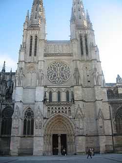A cathedral in white stone. The west façade has a single entrance with a monumental rose window above it. The entrance and rose window are flanked on each side by a gothic bell tower and spire.