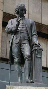 Statue of a man with a mortar and pestle in his left hand and his right hand upraised, holding a lens