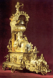 A regal, gold-coloured clock in the shape of an elephant-drawn chariot. Anthropomorphic dogs and other figures stand around him blowing trumpets and playing other instruments. The clock face is on Gambrinus' large belly. A plate of food is perched atop his gut, and a beer barrel is balanced over his head.