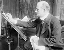 A man in late middle-age, facing left, drawing at an upright drawing board.