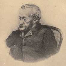 Black-and-white depiction from the waist up of an old, bearded man dressed in a dark jacket, sat in a chair and looking towards the left.