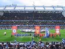 The Chelsea (blue) and Liverpool (red) teams line up side-by-side in front of banners bearing their club crests and the Champions League final logo. In the background, a flag bearing the Champions League logo is waved over the centre circle, while the main stand is further back, full of spectators. In the foreground, Chelsea fans wave blue flags bearing their club crest.