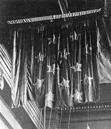 A black-and-white photo of a battle-worn flag suspended from the ceiling of a museum