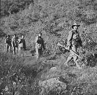 Three unarmed soldiers being led down a hill by two soldiers armed with rifles, one in the front the other in the rear.