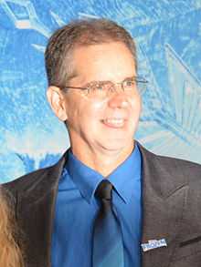 A man in his early fifties is standing in front of a blue wall. He is wearing a black coat over a blue collared shirt and a blue tie.