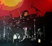 A man performing on a purple drum kit against a red-and-yellow backdrop. A drum stick is visible in his right hand; several microphones and a cooling fan are also visible.