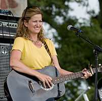 A woman wearing a yellow t-shirt smiling and playing a black acoustic guitar.