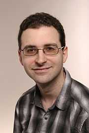 Head and shoulders picture of a clean-shaven young man with glasses and short brown hair, wearing a black and white plaid shirt that's open at the neck.