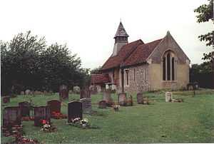 A small stone church with red tiled roofs seen through a churchyard from the southeast. On its far gable is a bellcote with a pyramidal roof