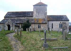 A long church with flint walls and tiled roofs.  A short tower with a pyramidal roof and two small windows rises slightly right of centre.  To the left, a longer section with a high roof, in front of which is a projection with a lower roofline and two low windows.  A porch with an arched doorway extends from the far left side.  To the right, there is a shorter and lower section with two windows.  A path leads through a graveyard to the porch.