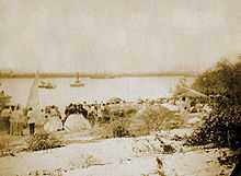 Photograph showing men and women in civilian dress lining a riverbank with small boats nearing the bank and large steamships in the distance near the opposite bank