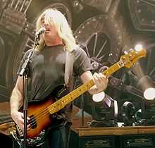 A fifty-eight-year-old man is singing into a microphone while playing a bass guitar. Behind him are stage lights and a partial view of a large steam train. His long white hair partly obscures his face. He wears a dark tee-shirt and darker pants.