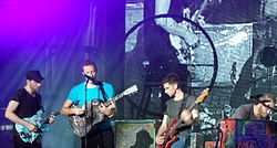 Three men play guitars, as one of them sings into a microphone. Another man looks downward, seated near a piano.