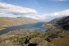 A big river and a nearby highway wind through a gorge between parallel ridges. The hills on the left are largely brown and treeless, while the hills on the right are partly covered by evergreen trees and grasses. A small lake lies in the foreground between the highway and the river.