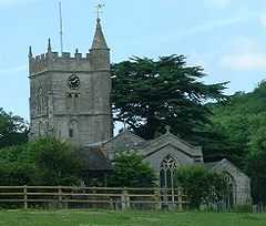 Gray stone building with arched windows. Square tower topped with spirelet, flagpole and weather vane. Foreground has small trees and bushes and a wooden rail fence.