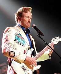 A man in a white jacket and blue shirt plays a white guitar.