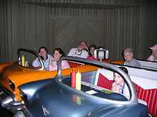 A photograph of seven people, including a girl, five women, and one man, all sitting in two booths designed to look like convertibles, one orange and one silver