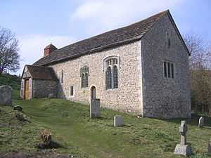 Three-quarter view of a long, low, flint rubble church on a rising grass slope with some gravestones. A tile-roofed porch juts out from the longer side, which also has two paired lancet windows and two other windows. The shorter side has three single-light round-headed windows and a blocked lancet window below the roofline.