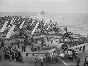 Black and white photo of a large number of single-engined monoplane aircraft on the deck of an aircraft carrier at sea. Another ship is visible in the background.