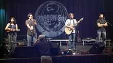 A woman and three men on stage; the woman with a ukulele and one of the men with a guitar.