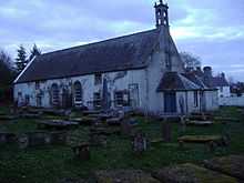 A T-plan kirk, limewashed whitewashed with a slate roof and small belfry. Many different-sized windows are visible.
