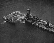 Aerial photo of a fireboat hosing down the stern of a battleship.