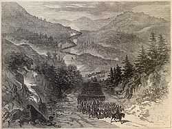 A lithograph in black and white of a road winding through a mountainous region.  An army of thousands is marching along the road, led by a military officer on a horse.