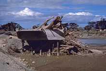 Image showing a mangled bridge split across a river with wood debris piled up against a section of the bridge.