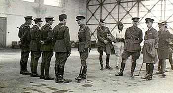 A group of military personnel in an aircraft hangar, four of whom are in a row facing another man, while the remainder stand informally, one of them wearing a flying suit