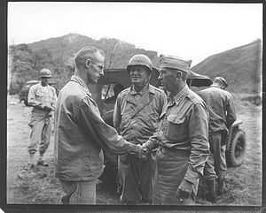 Two men in uniform shake hands. One is bear headed; Krueger wears his garrison cap. With them is a man wearing a steel helmet. A jeep is parked behind them.