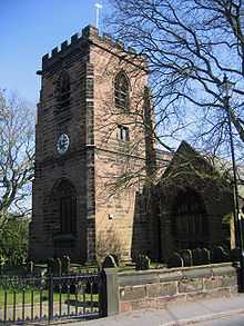 A square castellated tower viewed from one corner, with its front, incorporating a clock and an arched entrance, in shadow. The main church building, about half the height of the tower, is to the right and is connected to the tower.