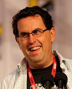 A man with black hair and glasses speaks into a microphone.