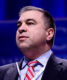 Head shot of dark haired young man with some five o'clock shadow, wearing blue shirt, red striped necktie, and dark jacket.