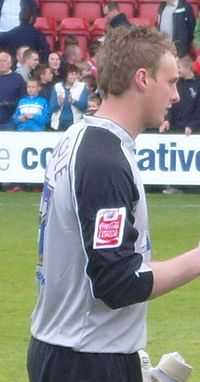 A man with brown hair is wearing a grey top and black shorts. He is standing on a grass field.