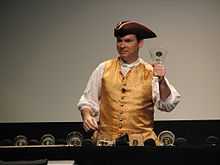 Man wearing tricorn hat and vest ringing a glass handbell