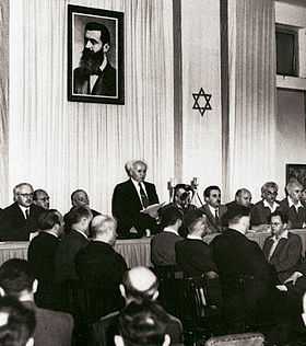 A man reads a document to a small audience assembled before him. Behind him are two elongated flags bearing the Star of David and portrait of a bearded man in his forties.
