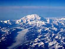 In an aerial image, a mountain is surrounded by many smaller mountains and a glacier.