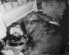 Middle-aged black-haired man lies face half-down on the floor, covered on his face and dark suit and trousers with blood. His hands are behind his back.