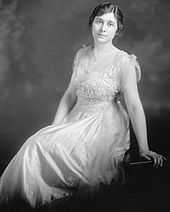 A slightly-built, dark haired woman wearing a white dress and leaning backward onto her left arm