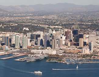 An overhead view of a San Diego's downtown with tall skyscrapers present throughout the area of different colors and sizes. A bay can be seen at the bottom of the image with several boats in the water. Also in the water is an aircraft carrier and a marina. In the background are other neighborhoods near the downtown area as well as mountains in the distance.