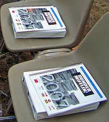 A book titled "Dunder Mifflin 2007 Annual Report" on top of a ream of paper in two empty plastic chairs